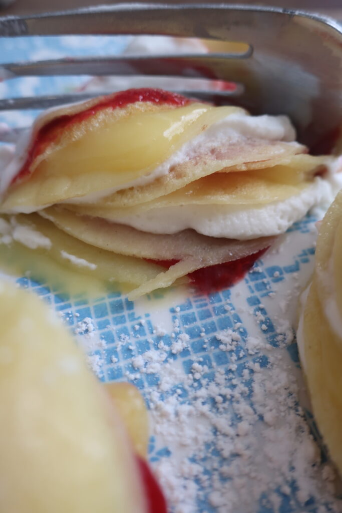 fork showing inside of mini crepes formed into stacks layered with homemade raspberry sauce, homemade lemon curd, and homemade whipped cream, and dusted with powdered sugar