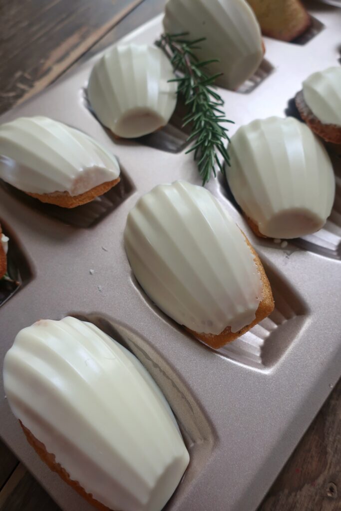 rosemary madeleines with a white chocolate shell in madeleine pan surrounded by fresh rosemary and striped blue napkin