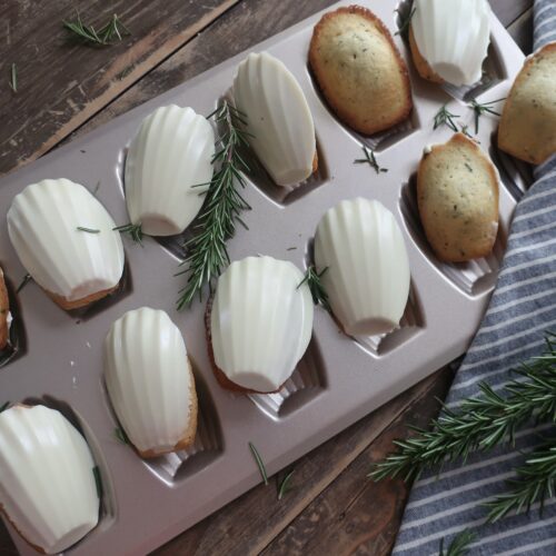 rosemary madeleines with a white chocolate shell in madeleine pan surrounded by fresh rosemary and striped blue napkin