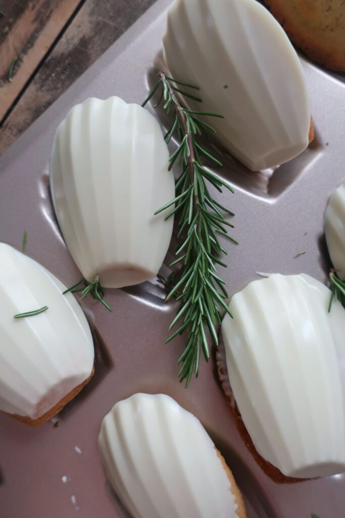 rosemary madeleines with a white chocolate shell in madeleine pan surrounded by fresh rosemary and striped blue napkin