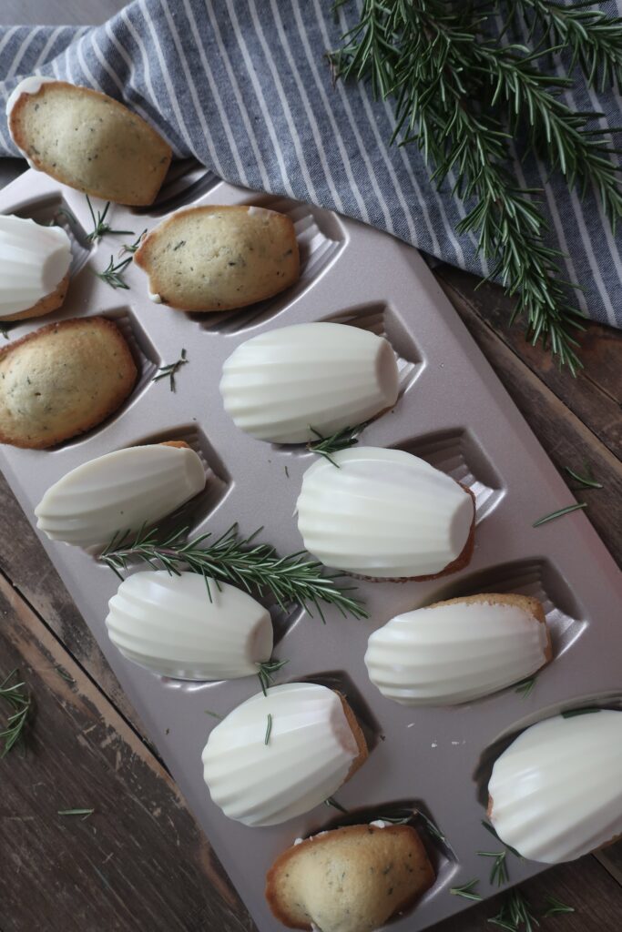 rosemary madeleines with a white chocolate shell in madeleine pan surrounded by fresh rosemary and striped blue napkin