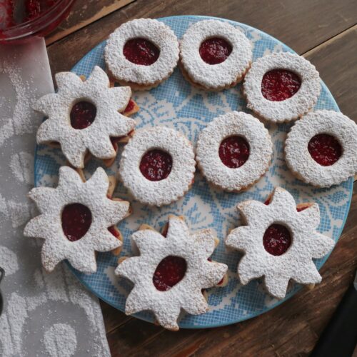 linzer cookie sandwiches with raspberry jam in the middle and dusted with powdered sugar on top; star and circle shaped linzer cookies on flowery blue plate