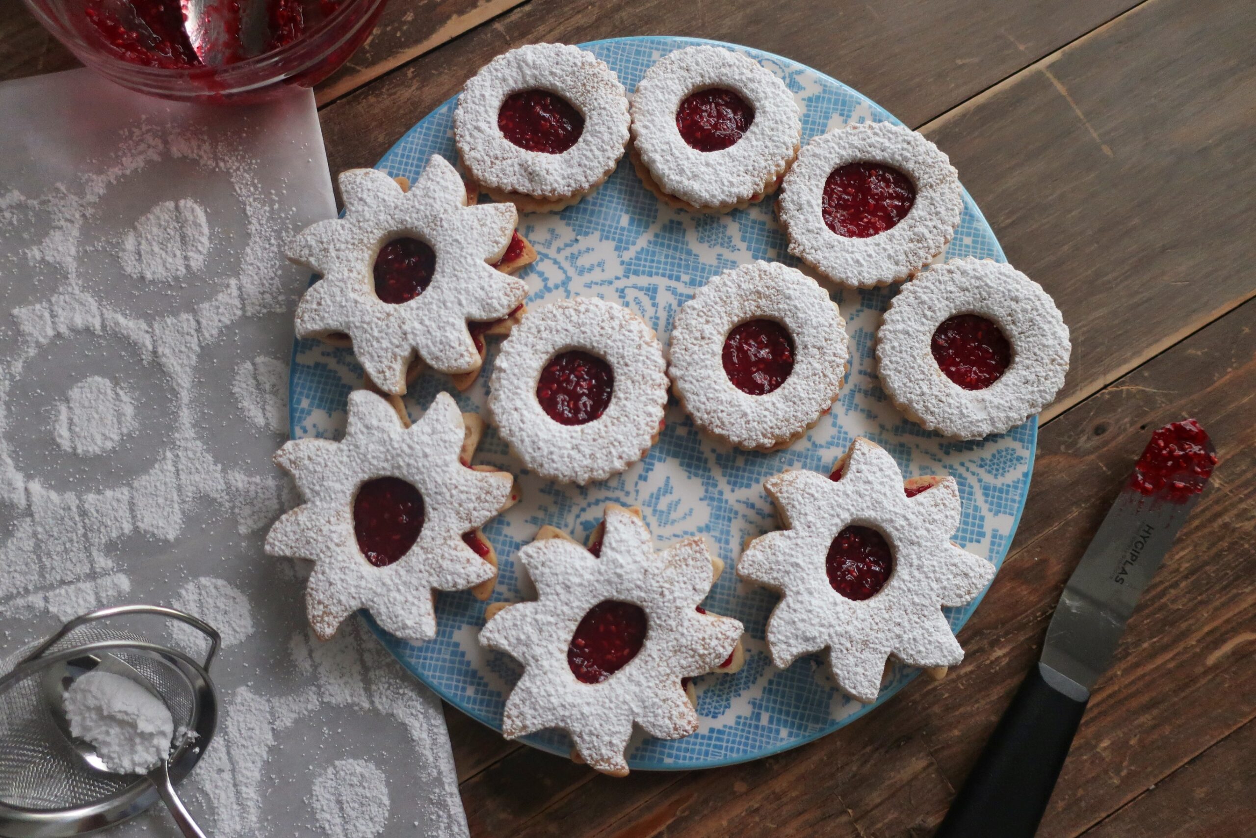 linzer cookie sandwiches with raspberry jam in the middle and dusted with powdered sugar on top; star and circle shaped linzer cookies on flowery blue plate