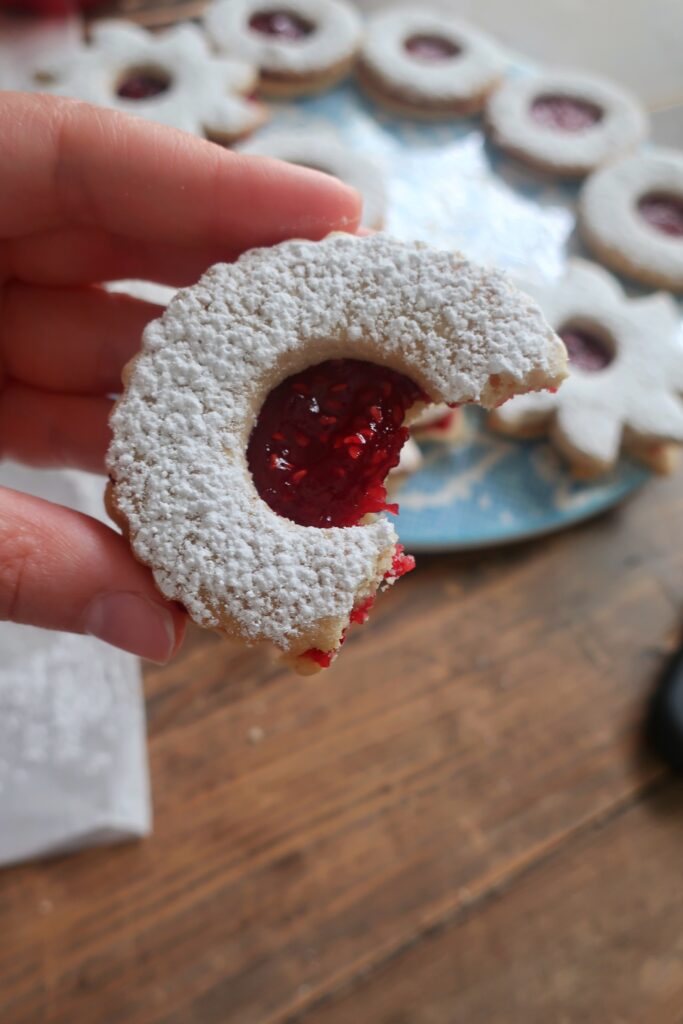 bite of a linzer cookie sandwiches with raspberry jam in the middle and dusted with powdered sugar on top; star and circle shaped linzer cookies on flowery blue plate