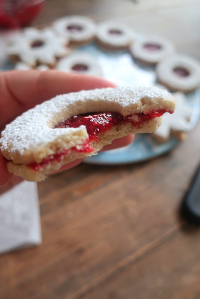 bite of a linzer cookie sandwiches with raspberry jam in the middle and dusted with powdered sugar on top; star and circle shaped linzer cookies on flowery blue plate