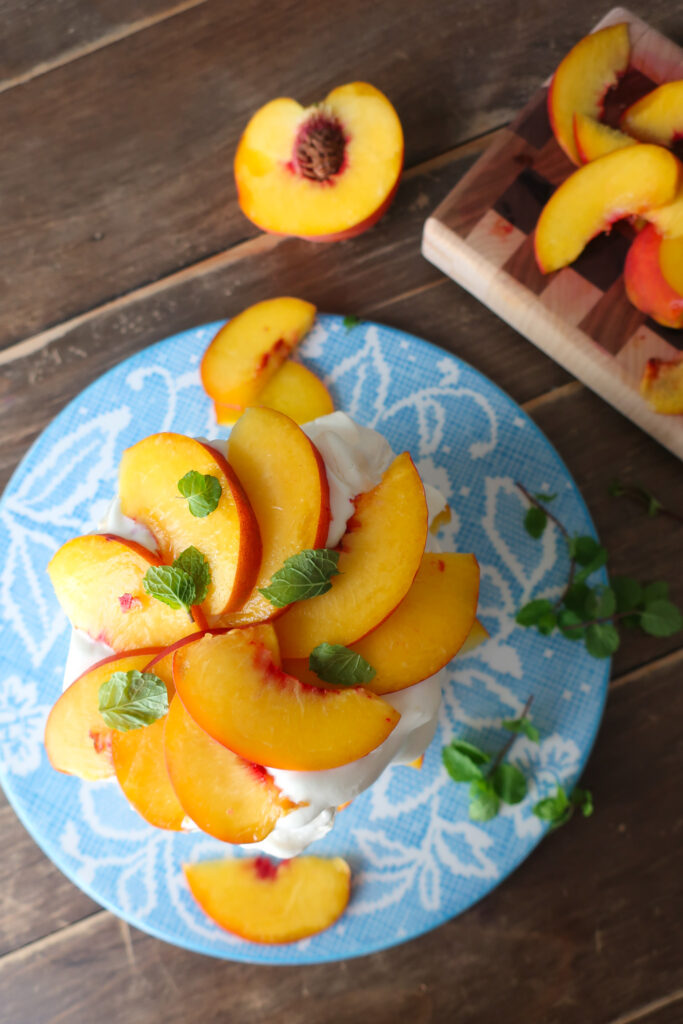 minty peach pavlova; two tiers of french meringue stacked with whipped cream, slices of fresh peaches, and chopped fresh mint