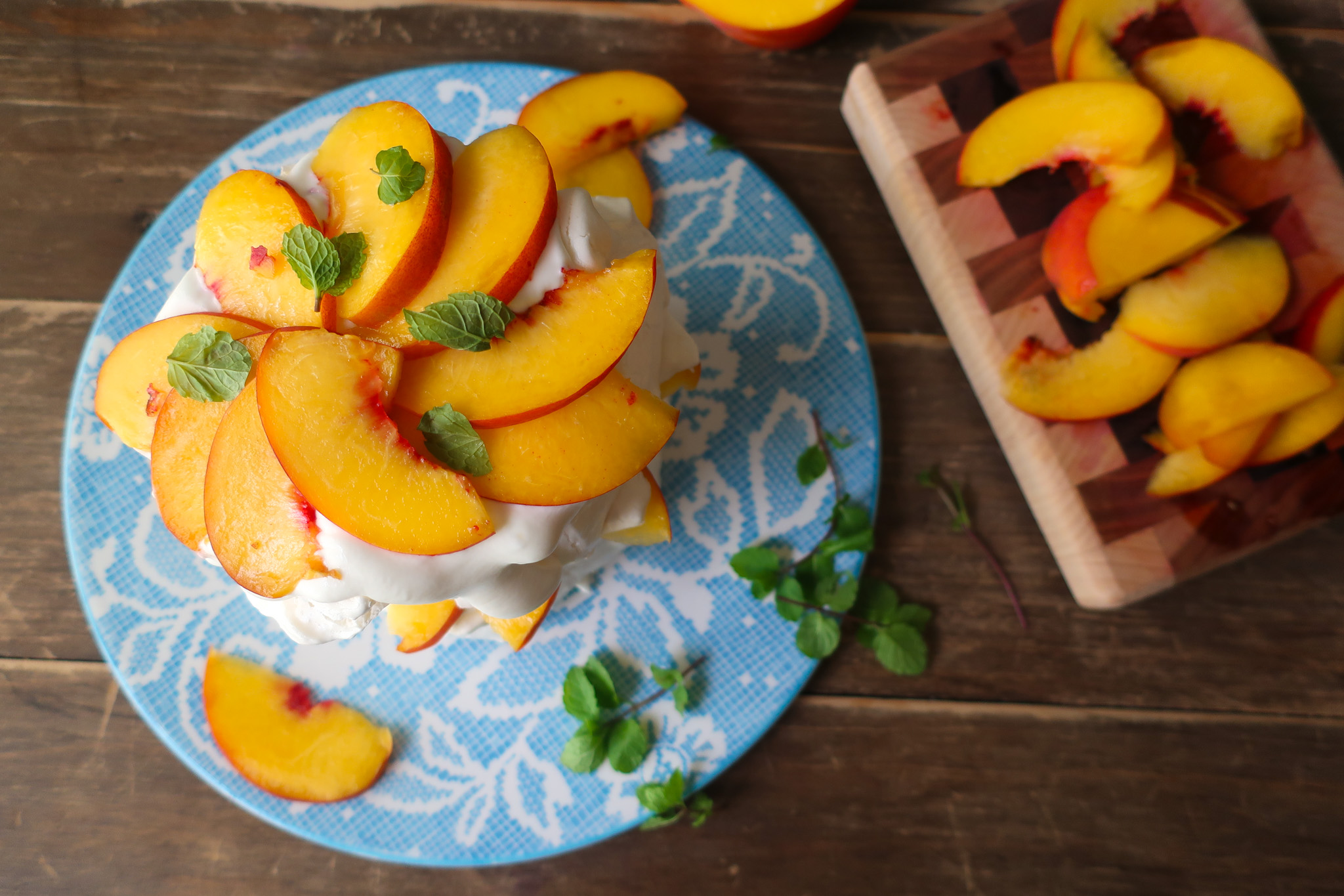 minty peach pavlova; two tiers of french meringue stacked with whipped cream, slices of fresh peaches, and chopped fresh mint