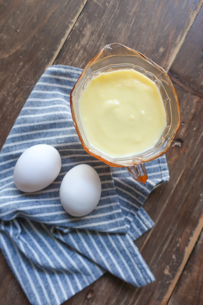 creme anglais in pale pink small pitcher sitting on blue striped napkin next to two eggs on wooden table