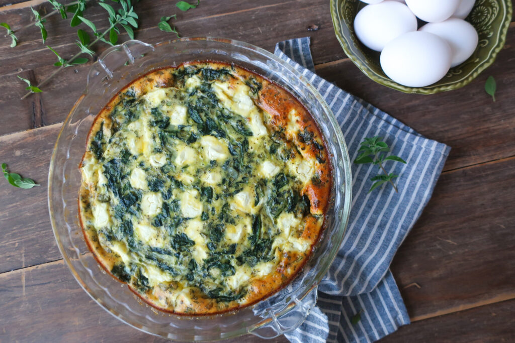 easy crustless mediterranean quiche with feta, spinach, oregano in pie dish, sitting on striped blue napkin next to bowl of eggs