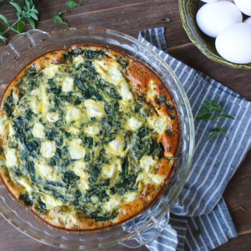 easy crustless mediterranean quiche with feta, spinach, oregano in pie dish, sitting on striped blue napkin next to bowl of eggs