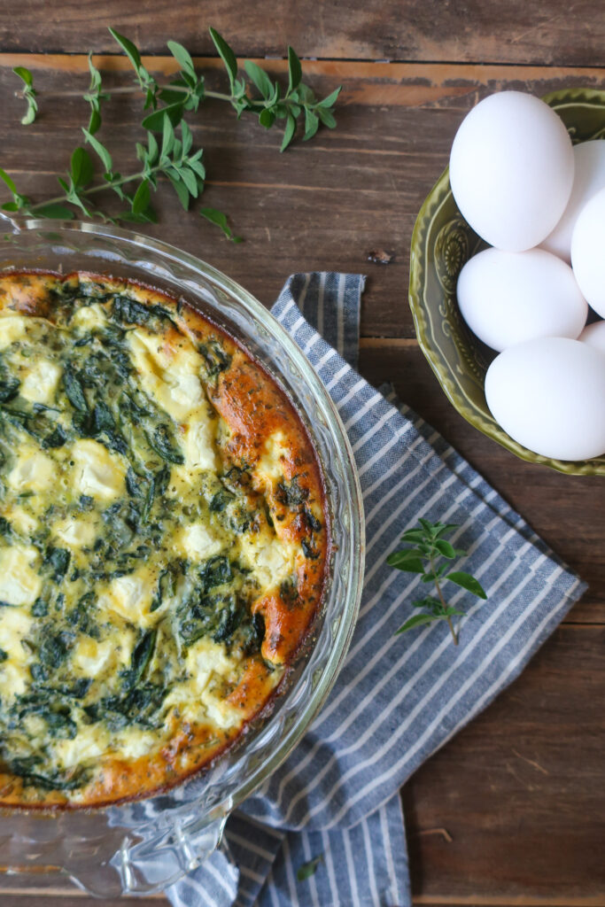 easy crustless mediterranean quiche with feta, spinach, oregano in pie dish, sitting on striped blue napkin next to bowl of eggs