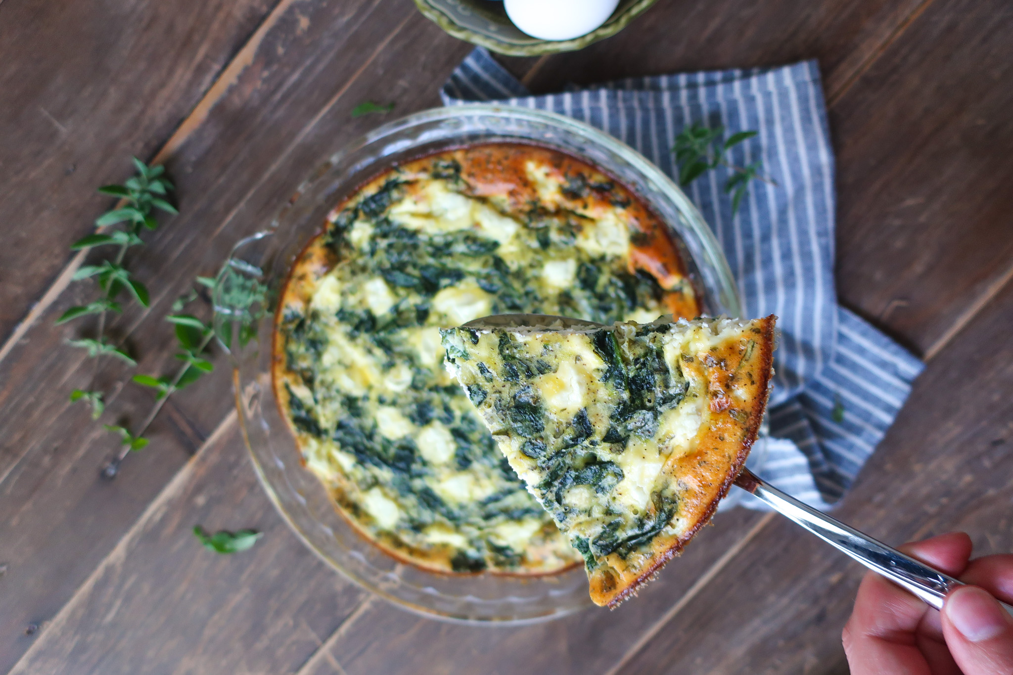 close up slice of easy crustless mediterranean quiche with feta, spinach, oregano in pie dish, sitting on striped blue napkin next to bowl of eggs