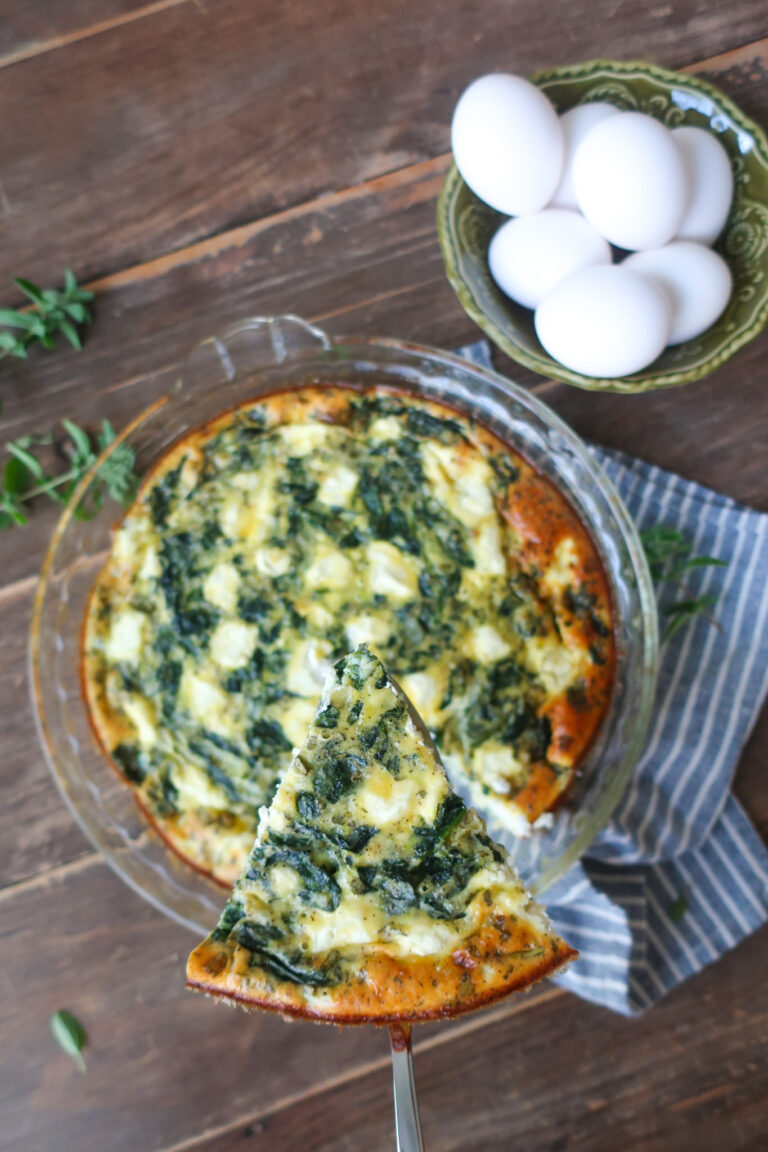 close up slice of easy crustless mediterranean quiche with feta, spinach, oregano in pie dish, sitting on striped blue napkin next to bowl of eggs