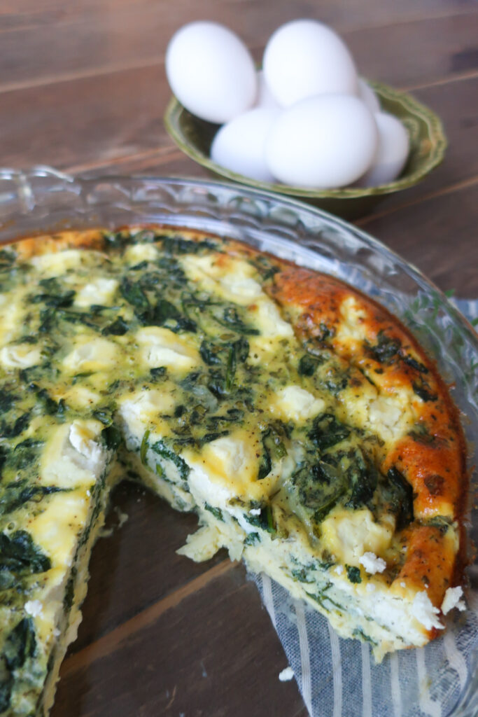 inside look at easy crustless mediterranean quiche with feta, spinach, oregano in pie dish, sitting on striped blue napkin next to bowl of eggs