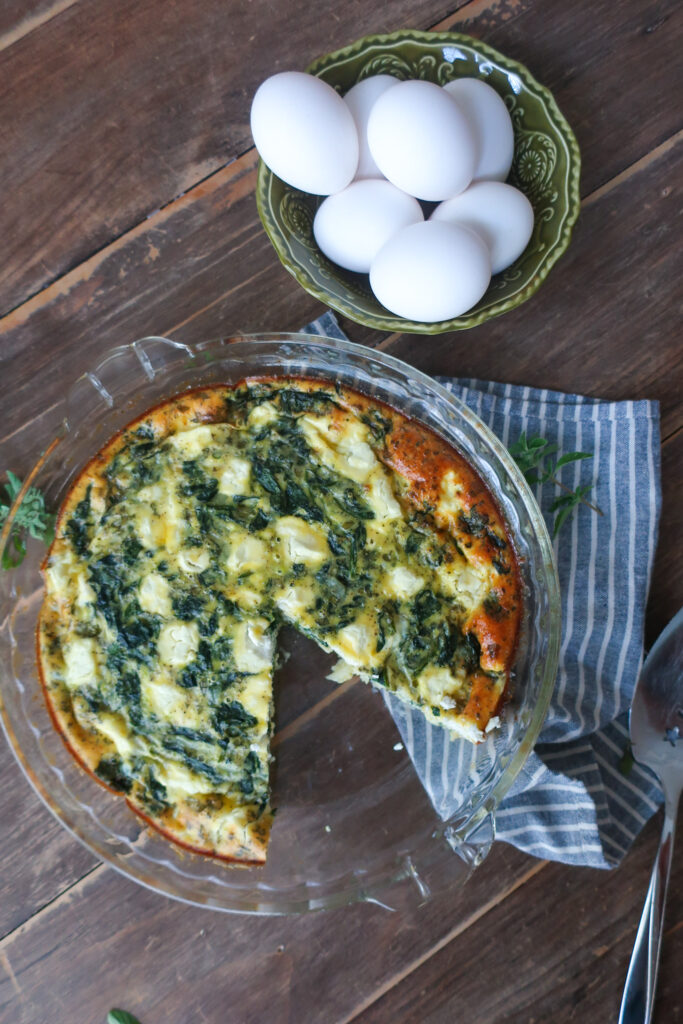 inside look at easy crustless mediterranean quiche with feta, spinach, oregano in pie dish, sitting on striped blue napkin next to bowl of eggs