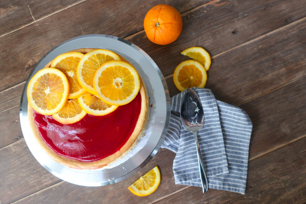 orange cheesecake with cinnamon spiced graham cracker crust and topped with cranberry sauce and sliced oranges; striped blue napkin next to orange slices and cheesecake