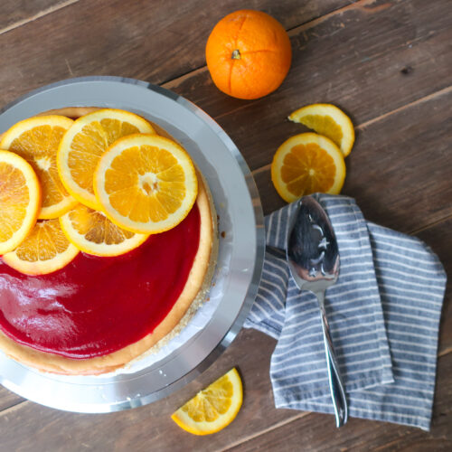 orange cheesecake with cinnamon spiced graham cracker crust and topped with cranberry sauce and sliced oranges; striped blue napkin next to orange slices and cheesecake