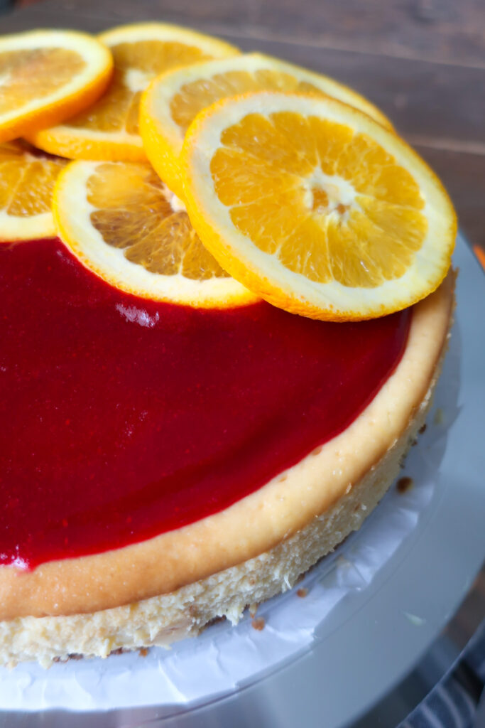 orange cheesecake with cinnamon spiced graham cracker crust and topped with cranberry sauce; orange slices on top of cheesecake topped with cranberry sauce