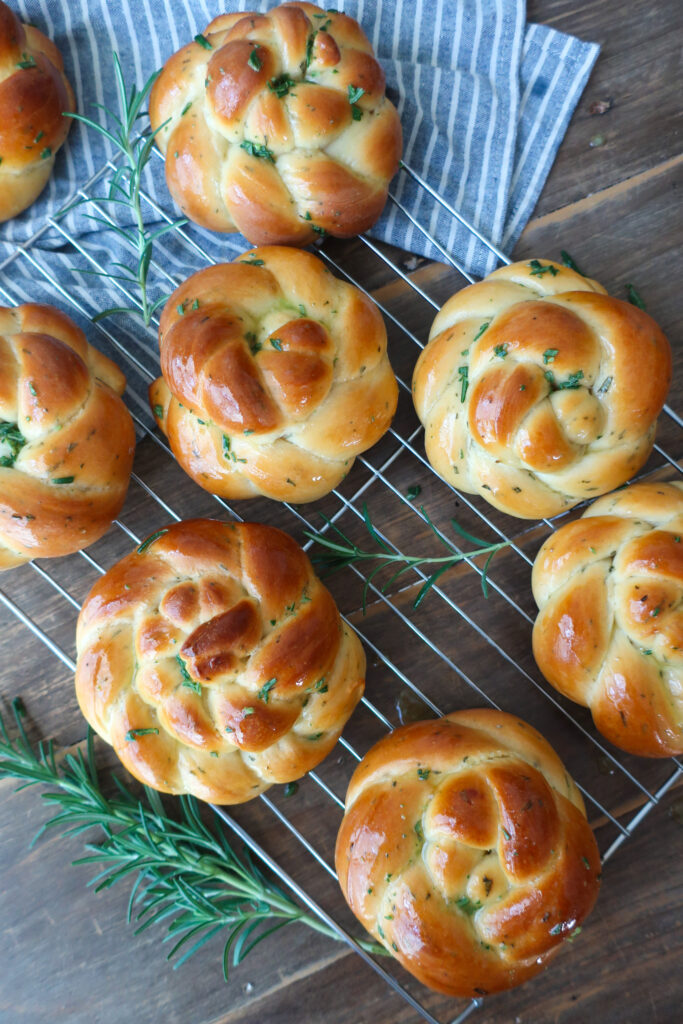 rosemary challah rolls baked and cooling on wire rack on top of blue striped napkin and surrounded by rosemary sprigs