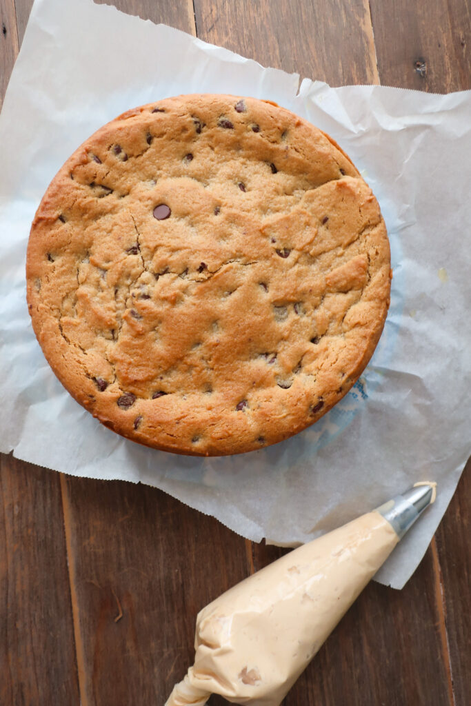 peanut butter and chocolate chip cookie cake before frosting with peanut butter buttercream frosting in piping bag next to it