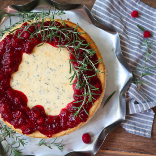 rosemary cheesecake with a cinnamon graham cracker crust base and topped with cranberry compote. decorate with fresh sprigs of rosemary sitting on a metal tray surrounded by a blue striped napkin, cranberries, and fresh rosemary