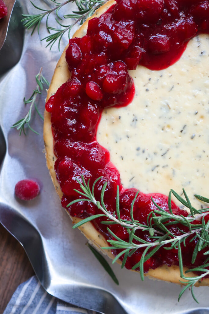 rosemary cheesecake with a cinnamon graham cracker crust base and topped with cranberry compote. decorate with fresh sprigs of rosemary sitting on a metal tray surrounded by a blue striped napkin, cranberries, and fresh rosemary