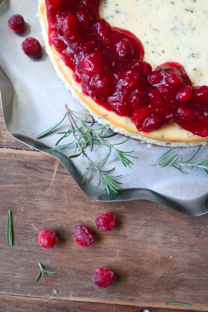 rosemary cheesecake with a cinnamon graham cracker crust base and topped with cranberry compote. decorate with fresh sprigs of rosemary sitting on a metal tray surrounded by a blue striped napkin, cranberries, and fresh rosemary