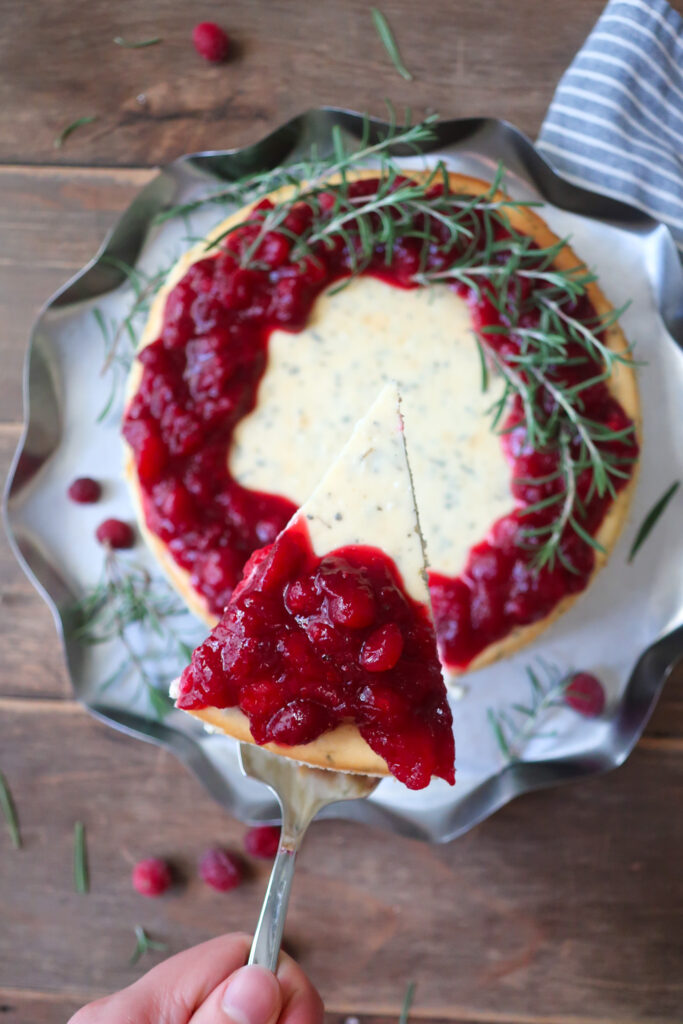 slice of rosemary cheesecake; rosemary cheesecake with a cinnamon graham cracker crust base and topped with cranberry compote. decorate with fresh sprigs of rosemary sitting on a metal tray surrounded by a blue striped napkin, cranberries, and fresh rosemary