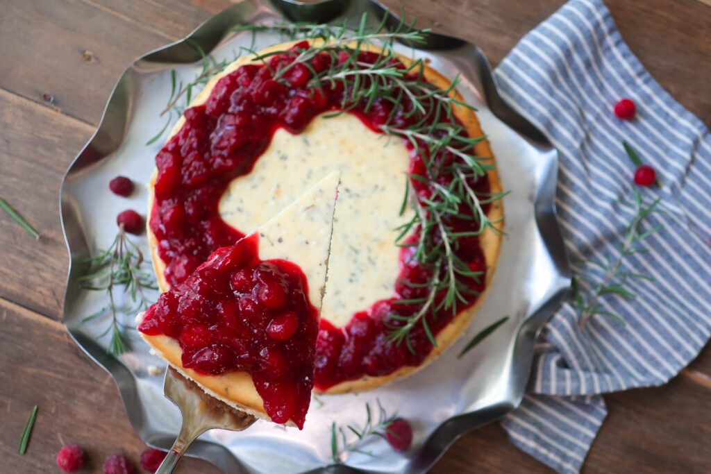 slice of rosemary cheesecake; rosemary cheesecake with a cinnamon graham cracker crust base and topped with cranberry compote. decorate with fresh sprigs of rosemary sitting on a metal tray surrounded by a blue striped napkin, cranberries, and fresh rosemary