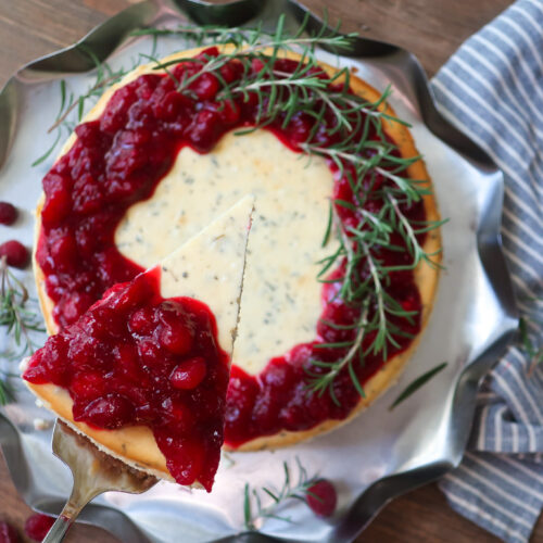 slice of rosemary cheesecake; rosemary cheesecake with a cinnamon graham cracker crust base and topped with cranberry compote. decorate with fresh sprigs of rosemary sitting on a metal tray surrounded by a blue striped napkin, cranberries, and fresh rosemary