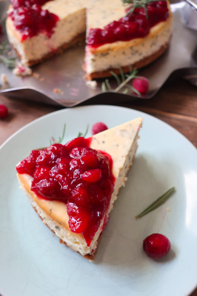 slice of rosemary cheesecake; rosemary cheesecake with a cinnamon graham cracker crust base and topped with cranberry compote. decorate with fresh sprigs of rosemary sitting on a metal tray surrounded by a blue striped napkin, cranberries, and fresh rosemary