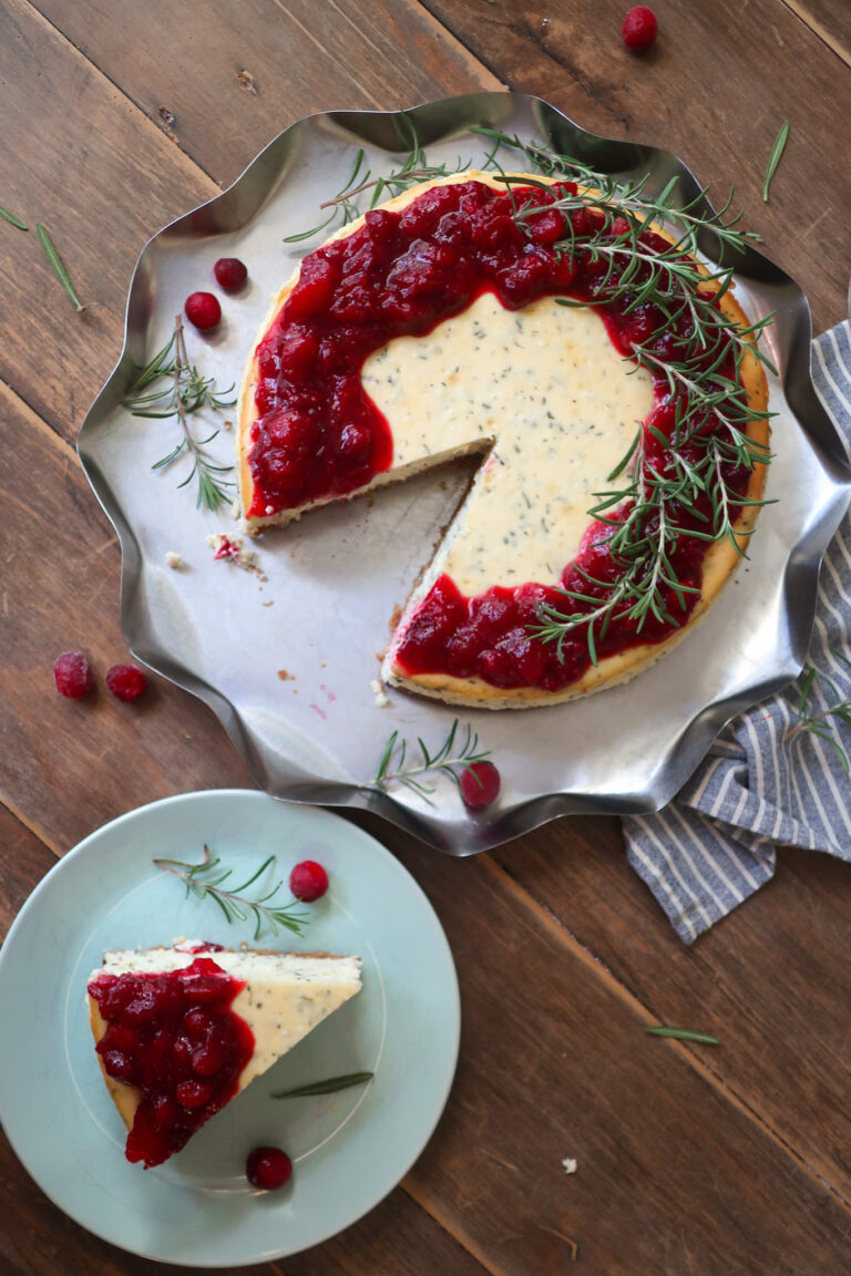 inside slice of rosemary cheesecake; rosemary cheesecake with a cinnamon graham cracker crust base and topped with cranberry compote. decorate with fresh sprigs of rosemary sitting on a metal tray surrounded by a blue striped napkin, cranberries, and fresh rosemary