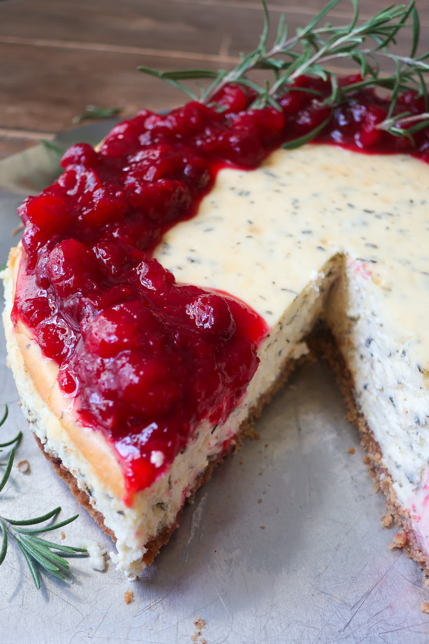 inside slice of rosemary cheesecake; rosemary cheesecake with a cinnamon graham cracker crust base and topped with cranberry compote. decorate with fresh sprigs of rosemary sitting on a metal tray surrounded by a blue striped napkin, cranberries, and fresh rosemary; rosemary cheesecake with cranberry compote and cinnamon spiced graham cracker crust