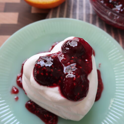 bowl of cherry berry compote on blue napkin with half of orange; baked heart-shaped pavlova made with french meringue filled with whipped cream and cherry berry compote