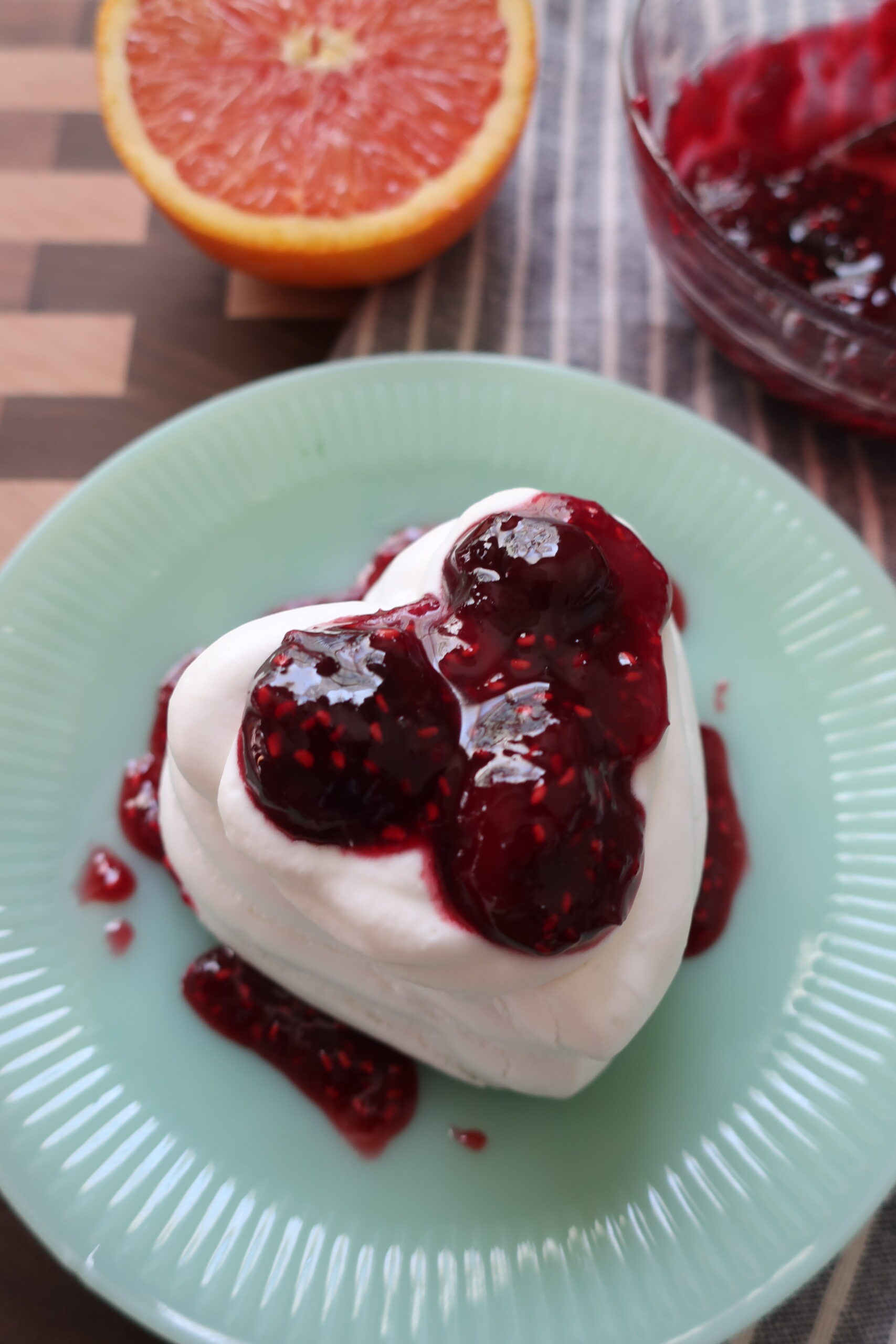 bowl of cherry berry compote on blue napkin with half of orange; baked heart-shaped pavlova made with french meringue filled with whipped cream and cherry berry compote