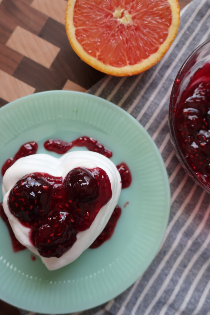 bowl of cherry berry compote on blue napkin with half of orange; baked heart-shaped pavlova made with french meringue filled with whipped cream and cherry berry compote