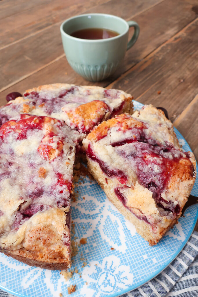 cherry coffee cake, coffee cake with cherry topping and streusel crumble, cherry coffee cake next to mug of hot drink, coffee cake next to mug of coffee