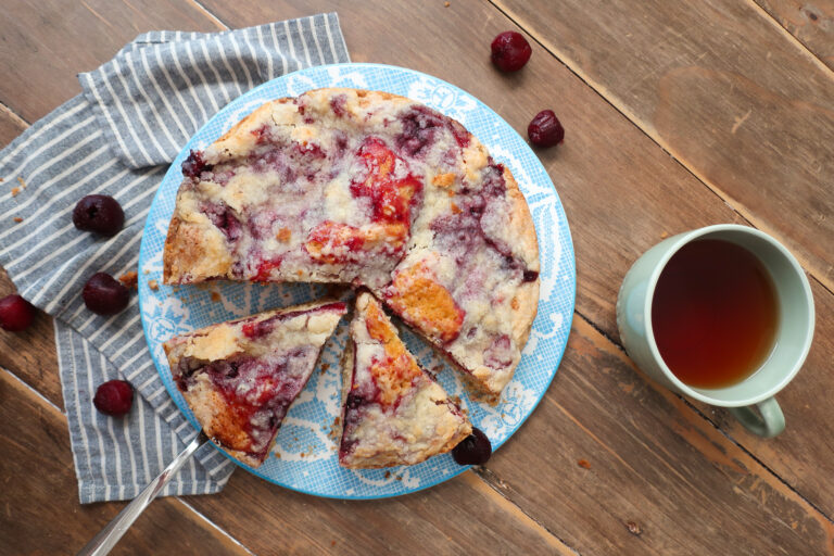 cherry coffee cake, coffee cake with cherry topping and streusel crumble, cherry coffee cake next to mug of hot drink, coffee cake next to mug of coffee