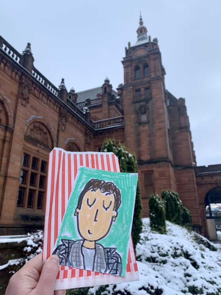 hand holding cartoon drawing of woman on a snowy day outside of the kelvingrove art gallery and museum in glasgow, scotland, uk, europe; solo travel; solo female travel; roadtrip scotland