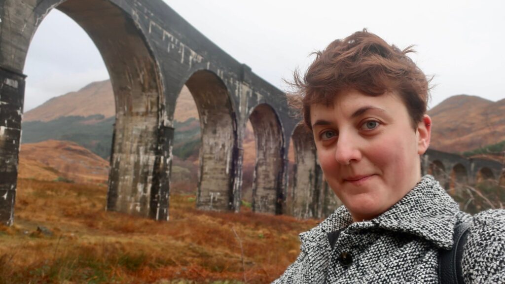 woman smirking near glenfinnan viaduct in scotland, united kingdom, europe; harry potter movies; solo travel; solo female travel; roadtrip scotland; roadtrip europe; scotland itinerary