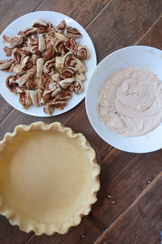 cut pieces of cinnamon rolls next to bowl of cream cheese filling next to crimped pie dough in pie dish for easy cinnamon roll pie with cream cheese filling