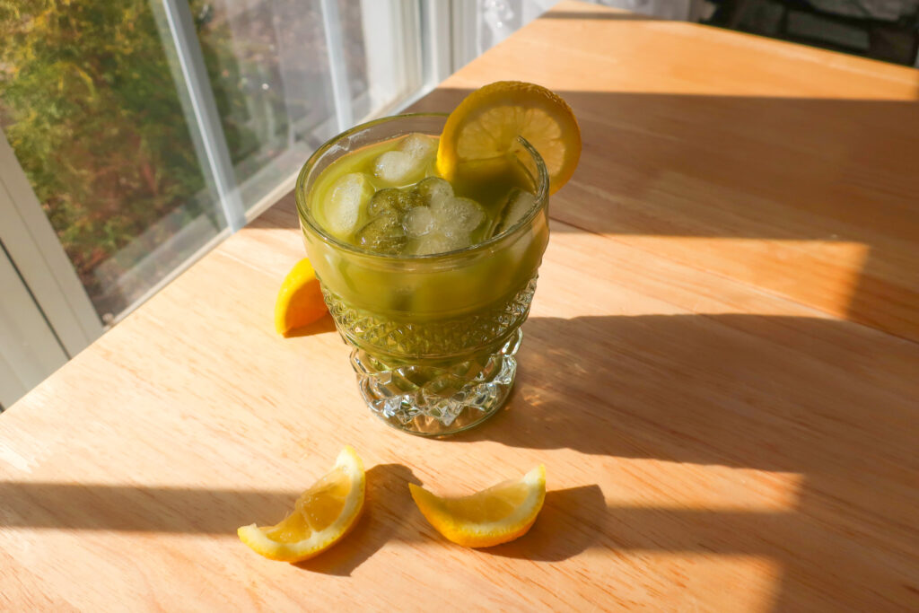 green drink in glass with lemon slice sitting in sunlight, easy iced matcha gingerade single-serve mocktail, ginger lemonade, st. patrick's day drinks, st. patrick's day mocktails, green drinks